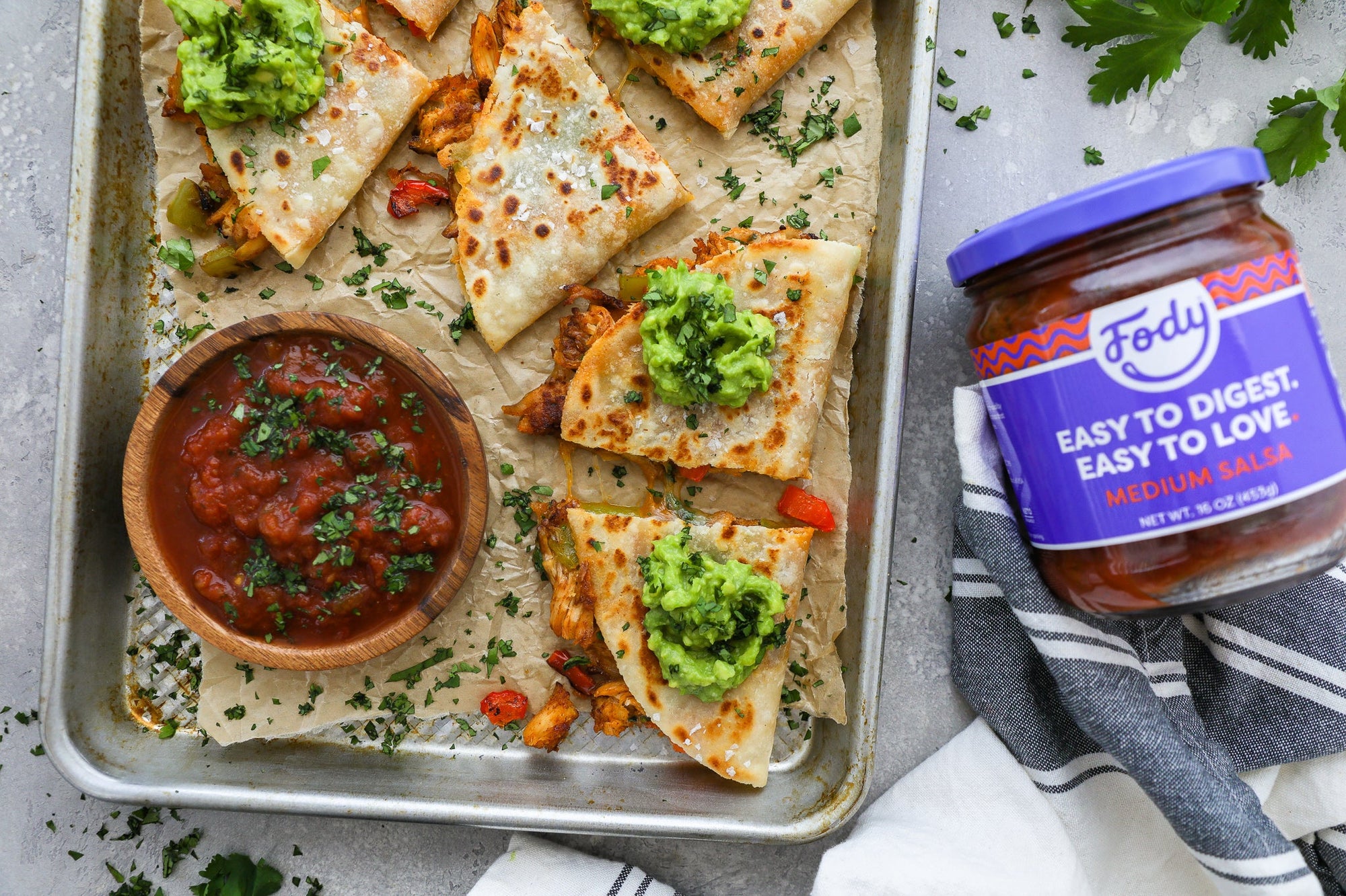 An image of Fody’s Seasoned Shredded Chicken Quesadillas. Low FODMAP quesadillas topped with guacamole sit on a baking tray beside a jar of salsa.