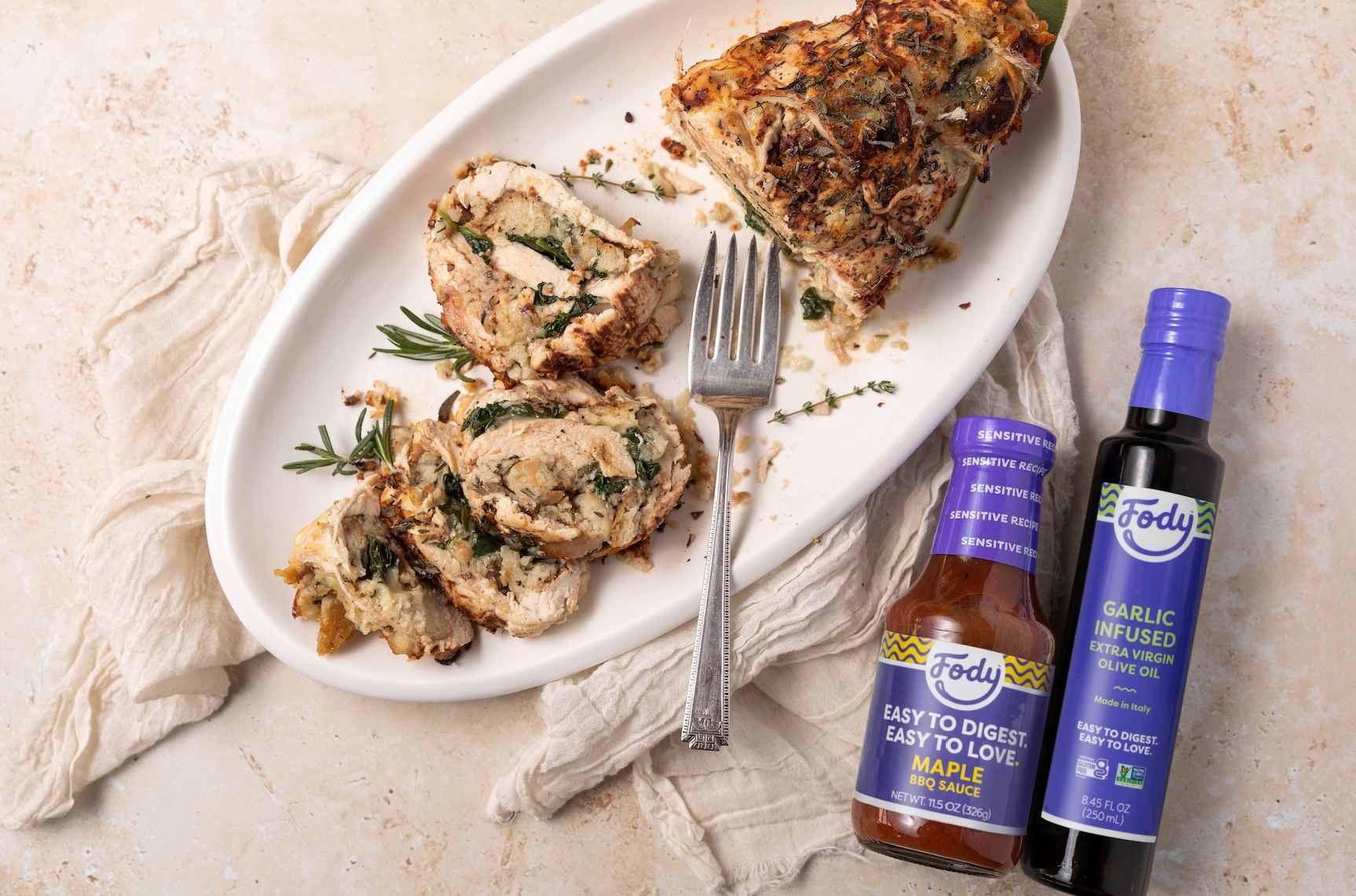 An image of Fody’s Spinach + Provolone Maple BBQ Turkey Roulade garnished with sprigs of rosemary, plated on an elegant white dish beside bottles of Fody's olive oil and BBQ sauce.