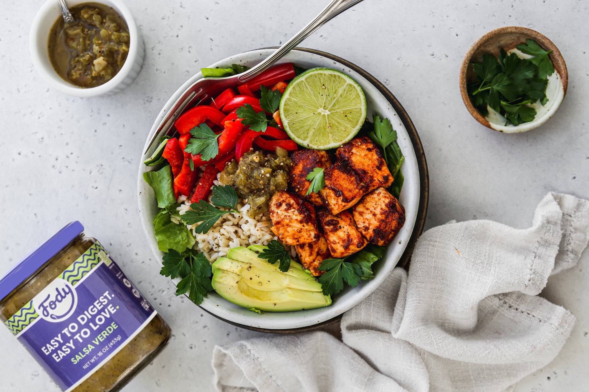 An image of Fody's taco salmon bites rice bowl laid out on a table beside a bottle of Fody's salsa verde and a white napkin.