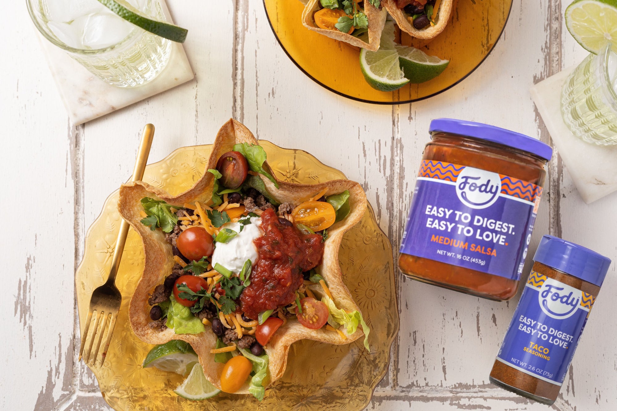 An image of Fody's Tortilla Bowl Taco Salad. A scalloped tortilla bowl filled with taco fillings sits on a rustic white wooden table, beside a mojito, a gold-coloured fork and two jars of Fody sauce and seasonings.