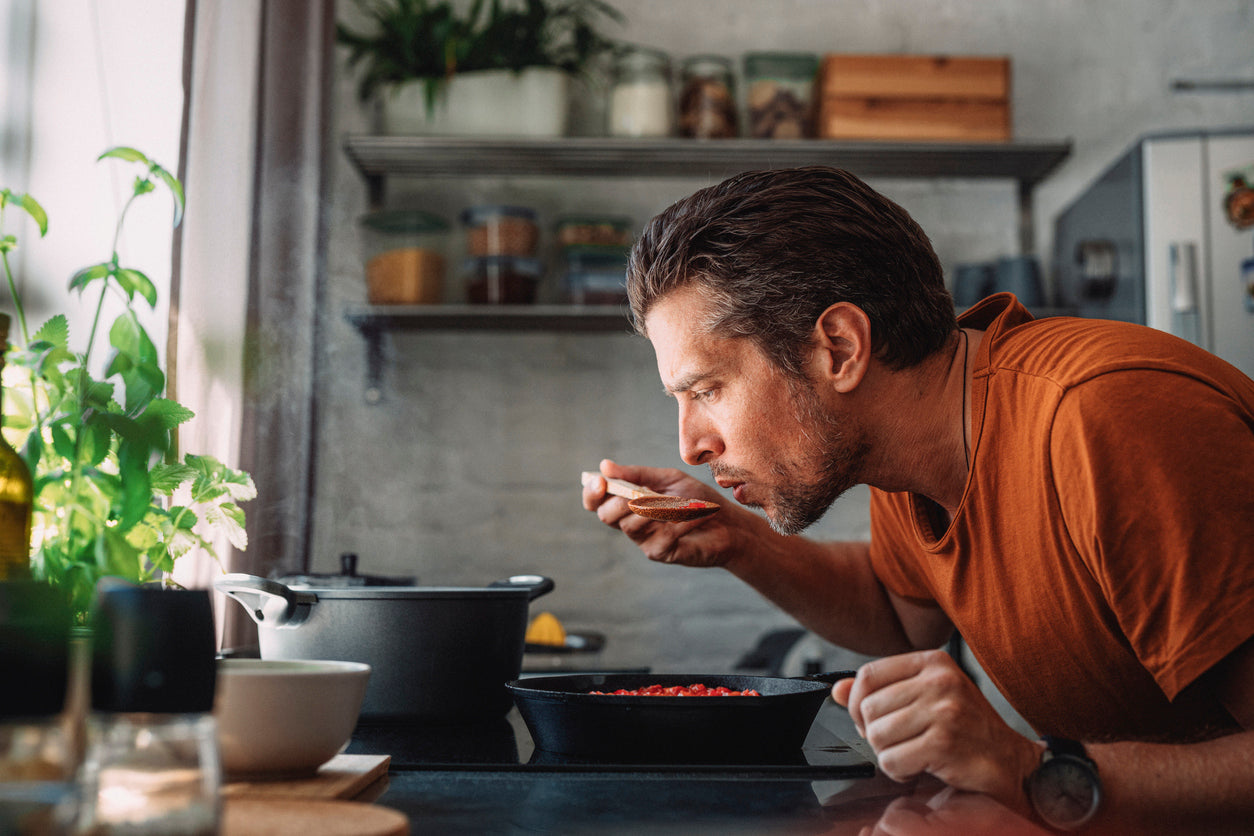 Healthy food that tastes good doesn’t have to be complicated! An image of a man at the stove, leaning towards a cast-iron pan to sample his cooking off a wooden spoon.