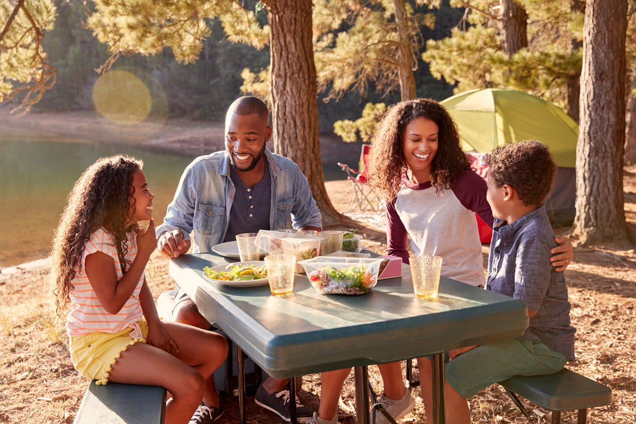 Gut friendly food ideas for picnics mean you can have a good time without worrying about where the bathrooms are! Image: a family sits around a picnic table at a campsite, enjoying homemade food on a sunny day.