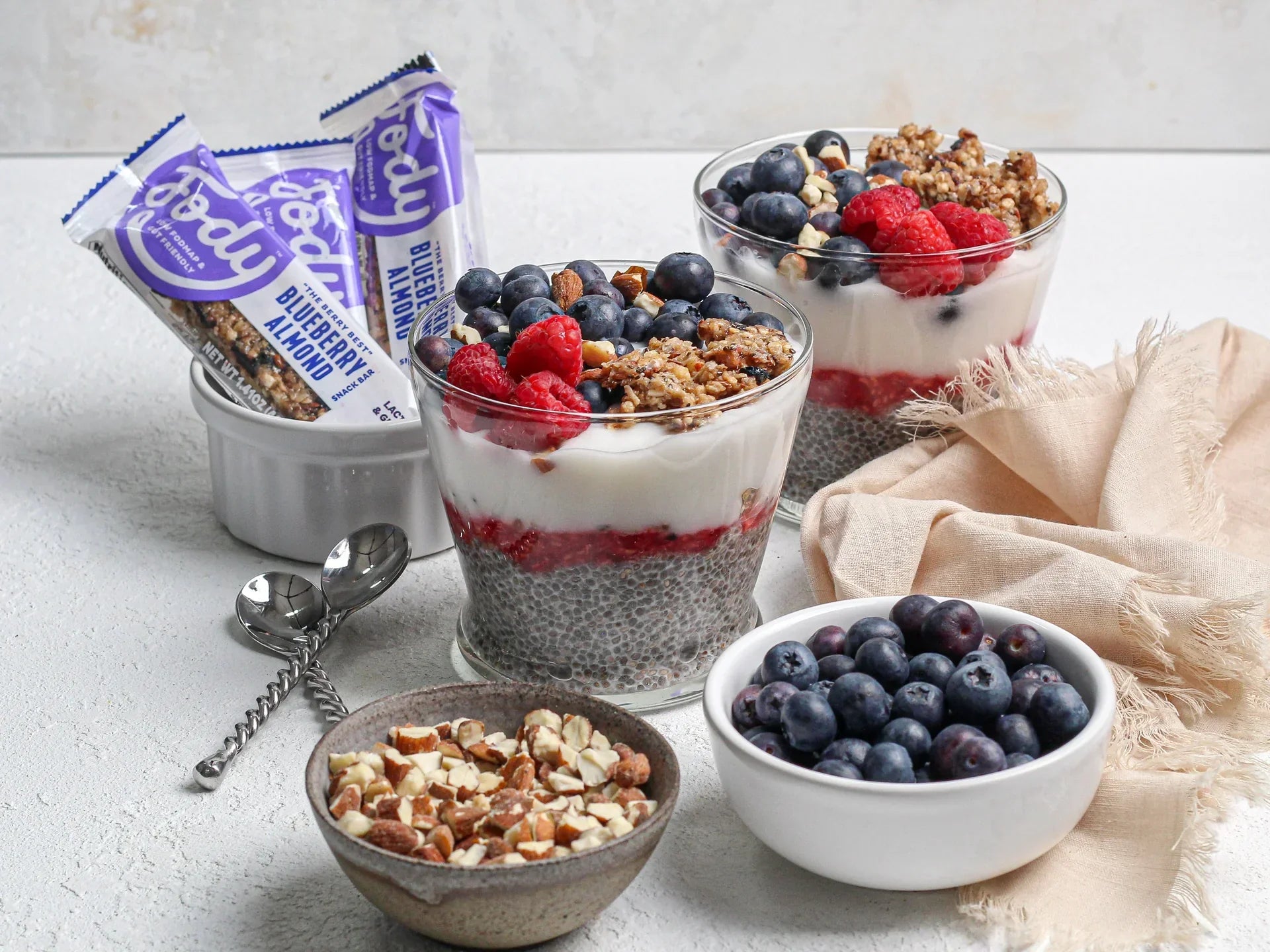 Fody Blueberry Almond Snack Bar beside chia pudding parfaits layered with yogurt, raspberries, and blueberries