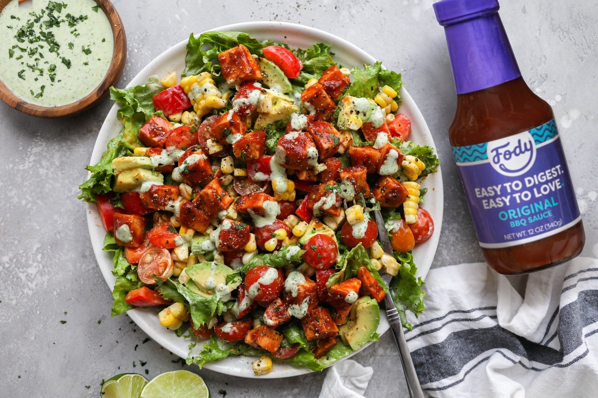 An image of Fody's BBQ chicken Cobb salad: a plate covered in corn, lettuce, bacon, cherry tomatoes, and BBQ chicken salad beside a bottle of Fody's maple BBQ sauce.