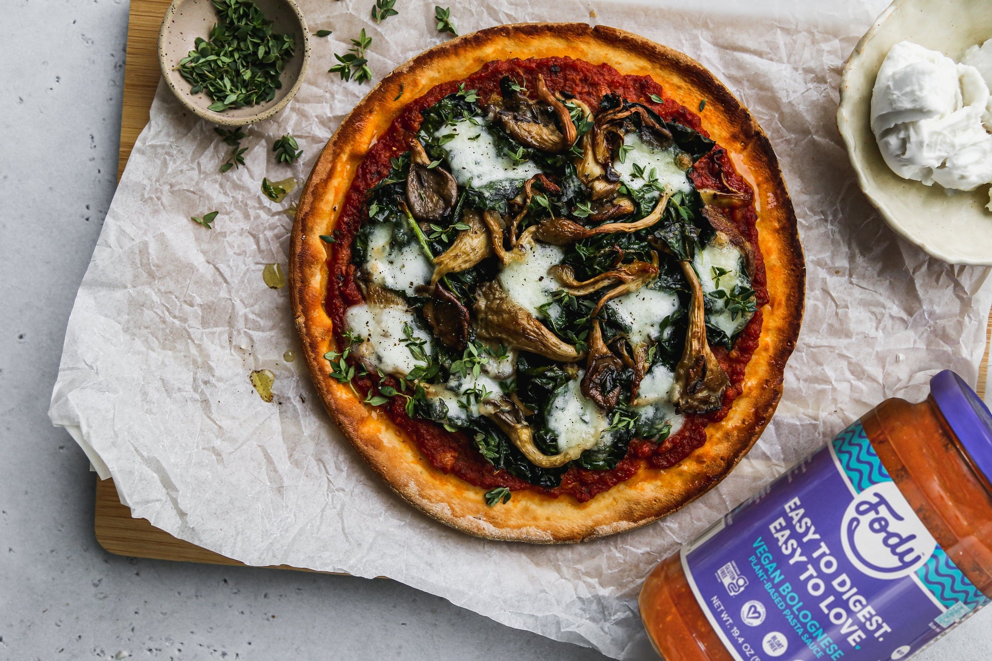 An image of Fody's Mushroom & Kale Bolognese Pizza. This low FODMAP pizza is on a sheet of parchment paper, beside a jar of Fody's vegan bolognese sauce.