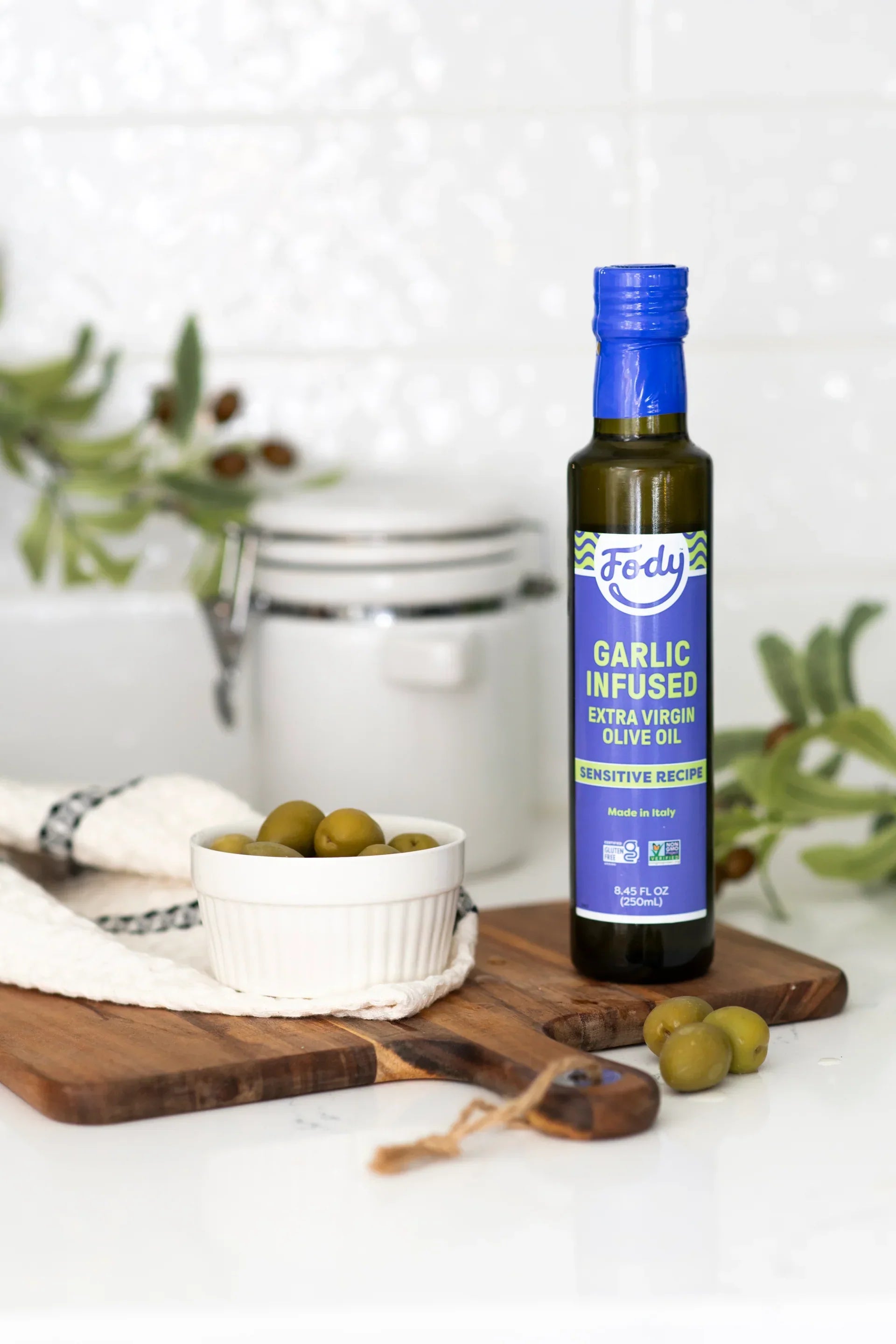 Fody Garlic Infused Extra Virgin Olive Oil 250ml bottle on a wooden cutting board with fresh green olives.