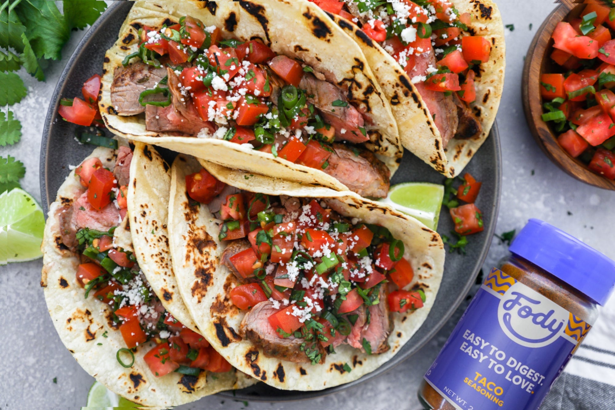 An image of Fody's grilled steak tacos with pico de gallo laid out on a plate beside a bottle of Fody's taco seasoning.