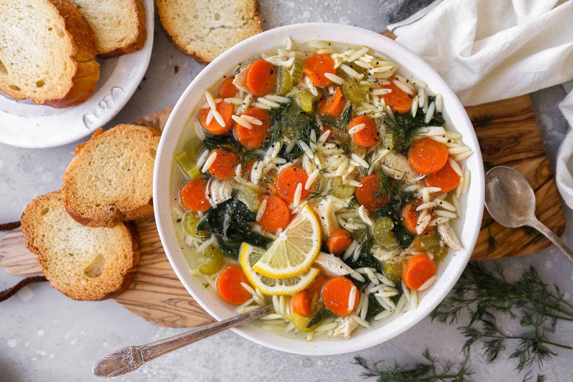 A bowl of Fody's Crockpot Lemon Chicken Orzo Soup sits on a table decorated with fresh herbs, white napkins, a wooden spoon and croutons. This low FODMAP chicken soup is in a white bowl, topped with lemon wedges.