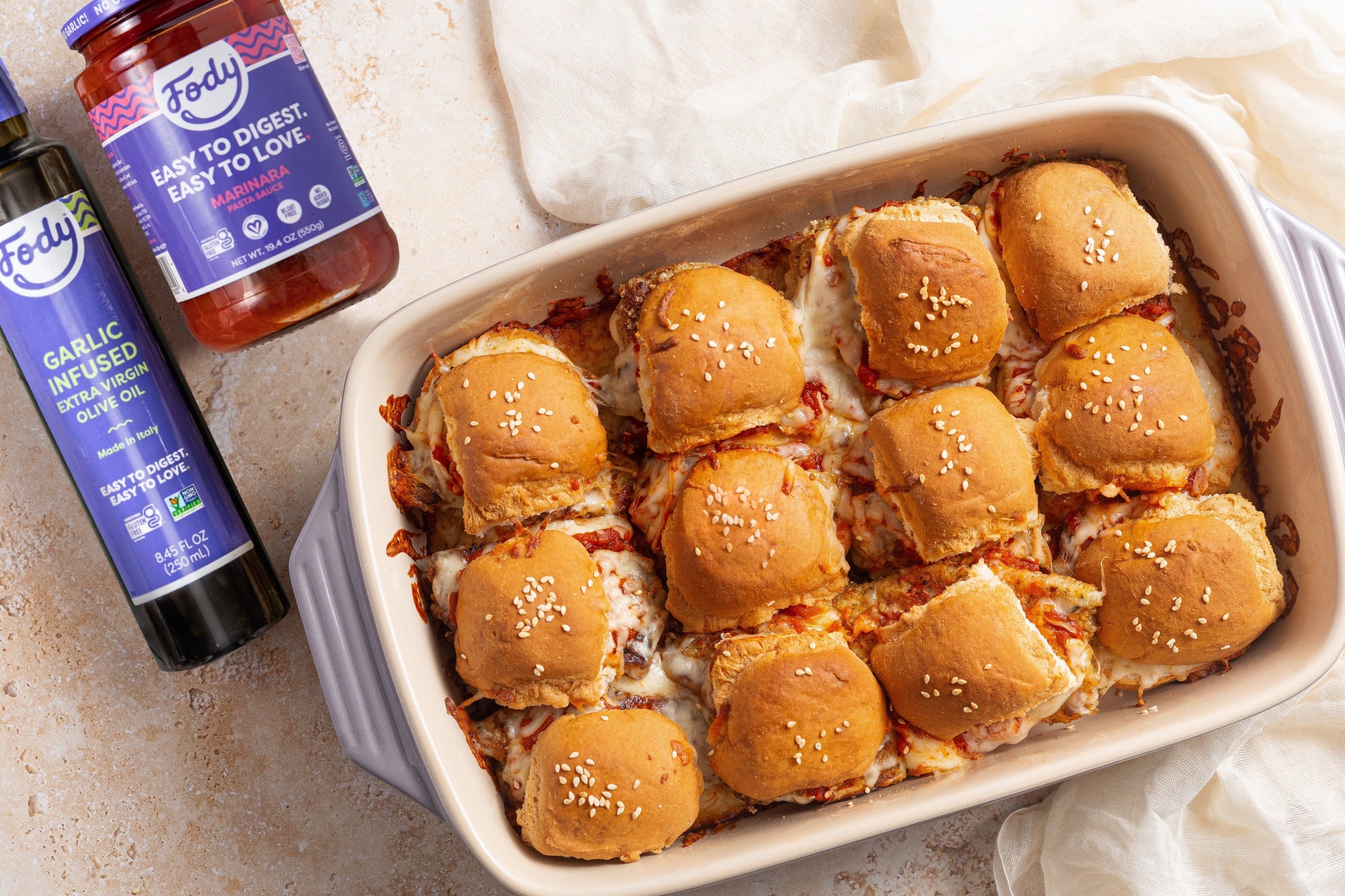 An image of Fody's chicken parm slider bake: a white baking pan of little chicken parm sliders beside a jar of low FODMAP marinara sauce and a bottle of low FODMAP olive oil.