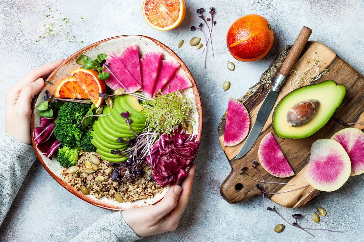 A plant-based diet doesn’t have to be complicated! Image: a pair of hands holding a colourful plate of grains, fruits and vegetables beside a cutting board covered in avocado and daikon radish.