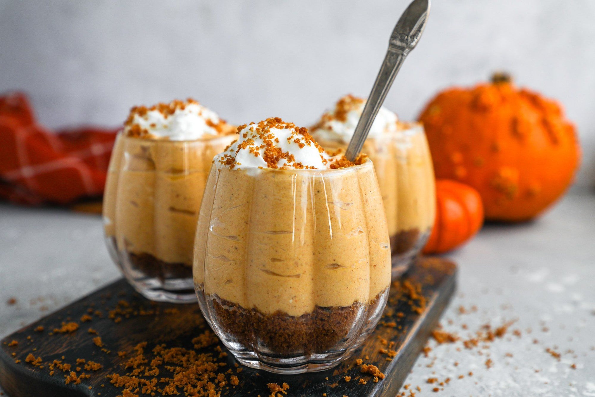 Three cups of Fody's No Bake Pumpkin Cheesecake Mousse sit on a cutting board sprinkled with decorative cinnamon. In the background behind the pumpkin cheesecake, three orange gourds decorate this image of a low FODMAP dessert.