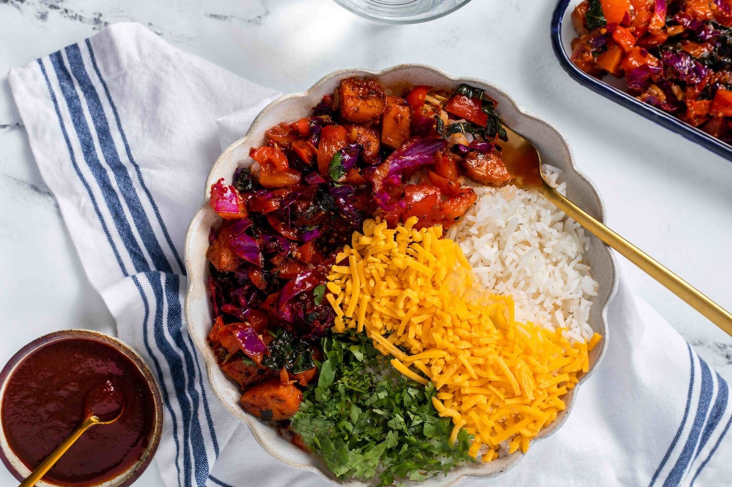 Image of a BBQ Buddha Bowl With Sweet Potato. This healthy vegetarian dinner is in a bowl with scalloped edges and contains a variety of vegetables, shredded cheese and rice, as well as a gold-coloured spoon. The bowl rests on a blue and white napkin.