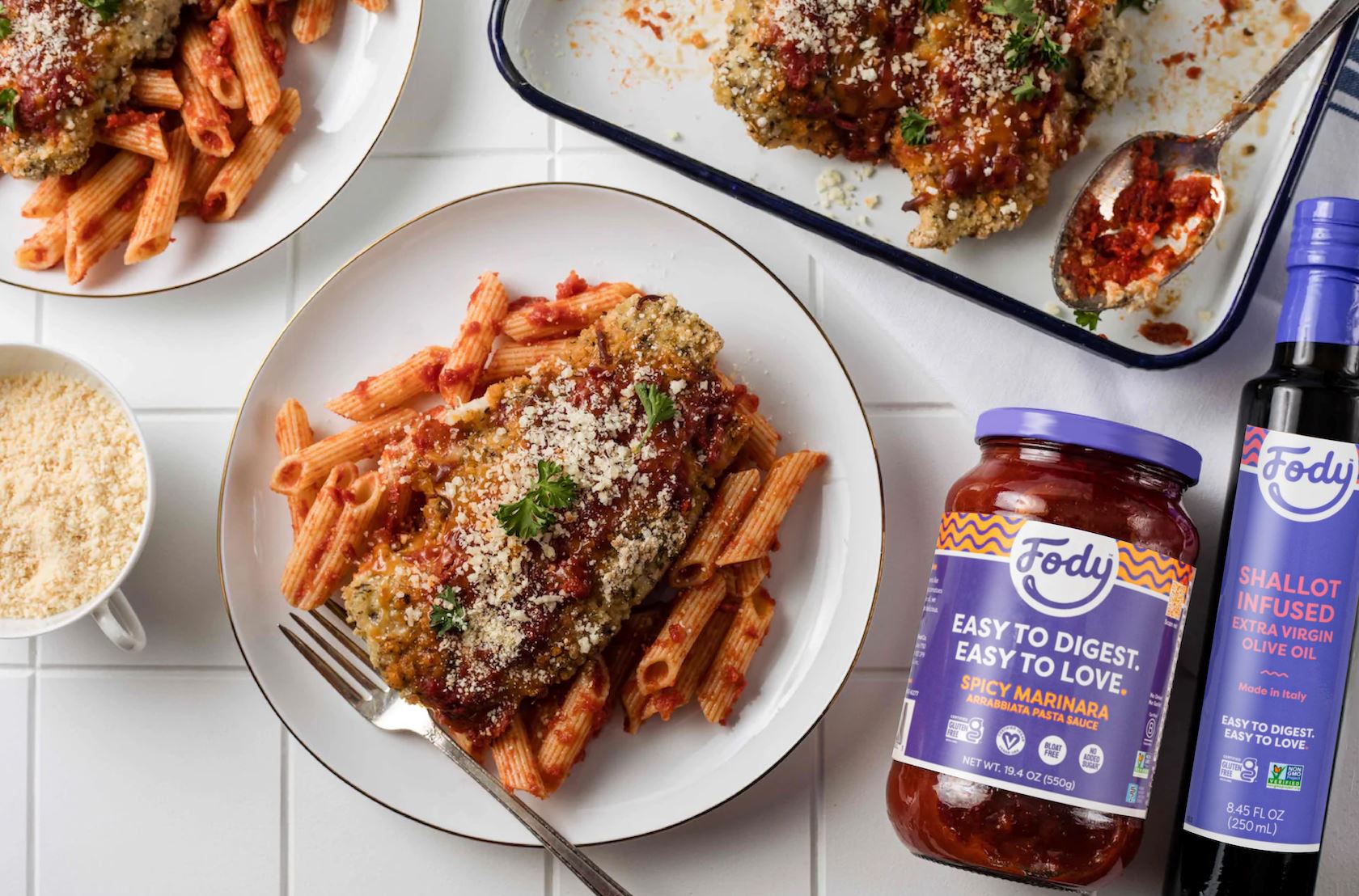 A plate loaded with Fody's chicken parmesan pasta bake with penne arrabiata. Beside the plate is a jar of Fody's Spicy Marinara sauce and a pinch bowl of parmesan. 