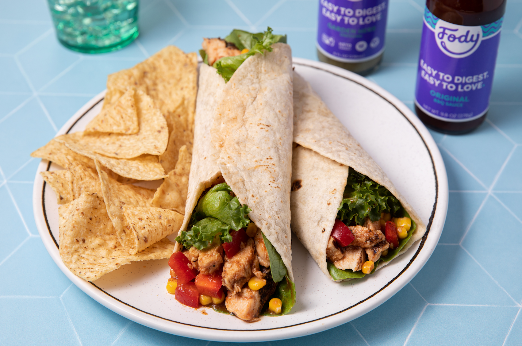An image of Fody's BBQ chicken salad wrap, on a plate beside chips and a bottle of Fody's Garden Herb sauce.
