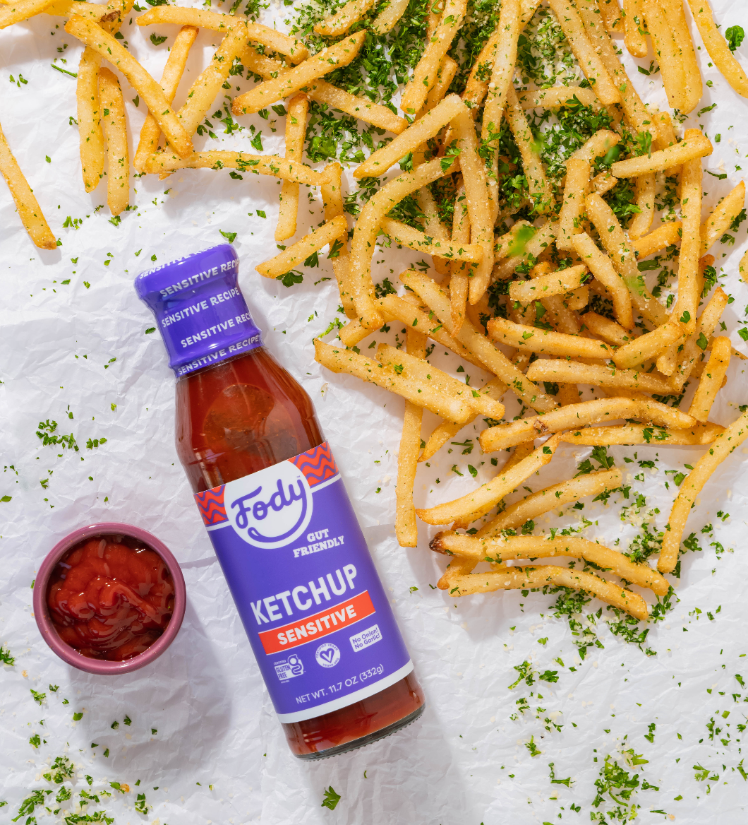 Crispy French fries sprinkled with herbs beside a bottle of ketchup and a small bowl of sauce on a white crinkled surface.