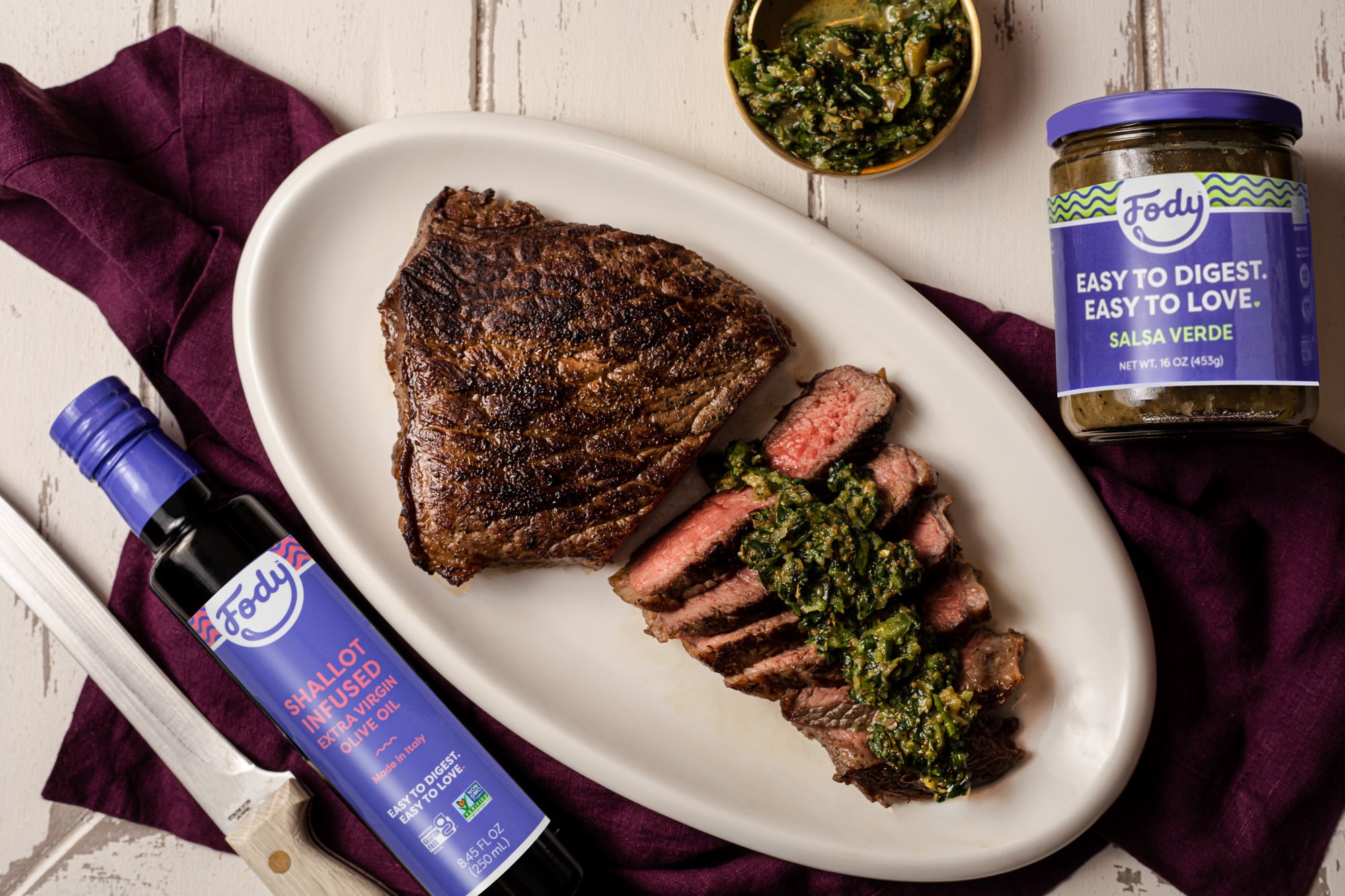 An image of Fody's steak with salsa verde chimichurri sauce. The steak is finely cut and topped with the green garnish. Bottles of Fody Foods salsa verde and olive oil are displayed alongside the steak dinner.
