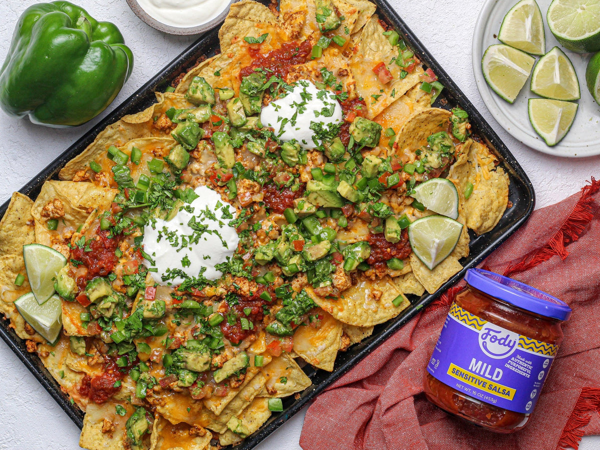 Low FODMAP nachos on a sheet pan topped with salsa, avocado, jalapeno, cilantro and lime wedges, beside a jar of Fody salsa.