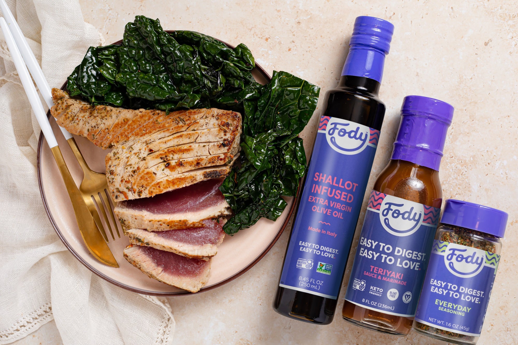 An image of Fody's Seared Teriyaki Tuna Steaks with Sauteed Kale, arranged on a plate next to elegant silverware and three bottles of Fody's seasonings. More than just tuna and kale! 