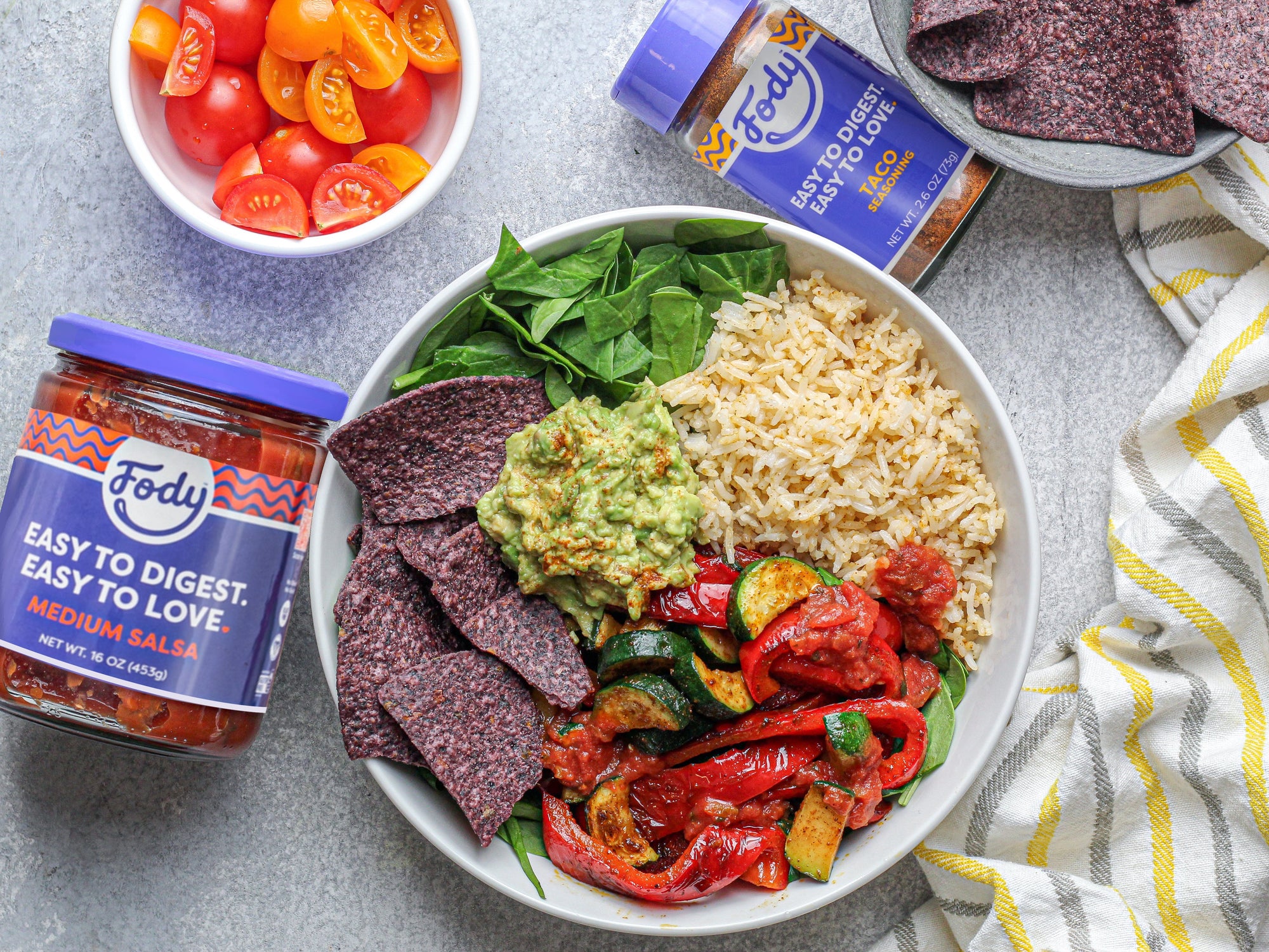 An image of Fody's bloat-free veggie burrito bowl: a white bowl filled with rice, corn chips, peppers, spinach, and tomato beside a jar of Fody's salsa and a jar of Fody's taco seasoning.