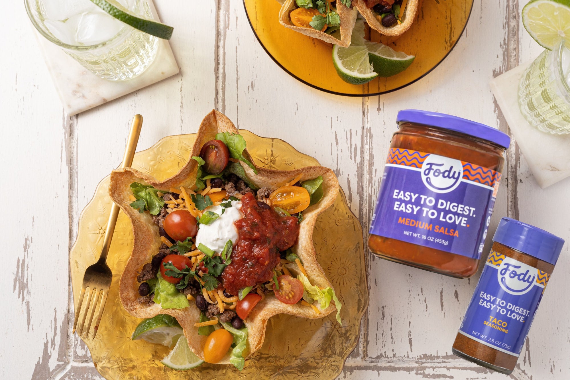 An image of Fody's Tortilla Bowl Taco Salad. A scalloped tortilla bowl filled with taco fillings sits on a rustic white wooden table, beside a mojito, a gold-coloured fork and two jars of Fody sauce and seasonings. 