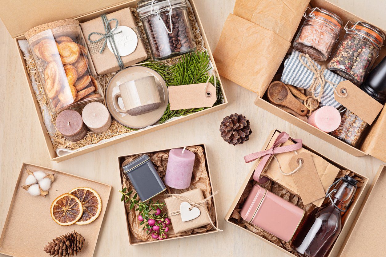 An image of a selection of consumable gift ideas in brown wrapping paper. Four boxes of healthy consumable gifts, including jars of peppercorns, dried oranges, candles, tins of spices and maple syrup. The table is decorated with pinecones.