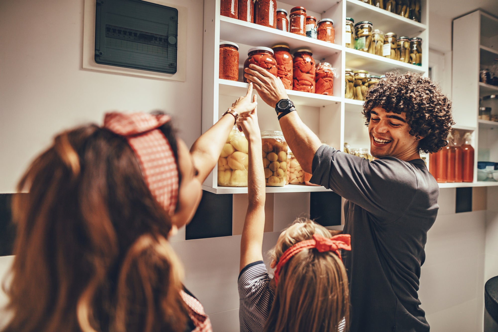 A family preparing to cook together. A father reaches for a jar among his pantry staples, smiling at his daughter and partner.