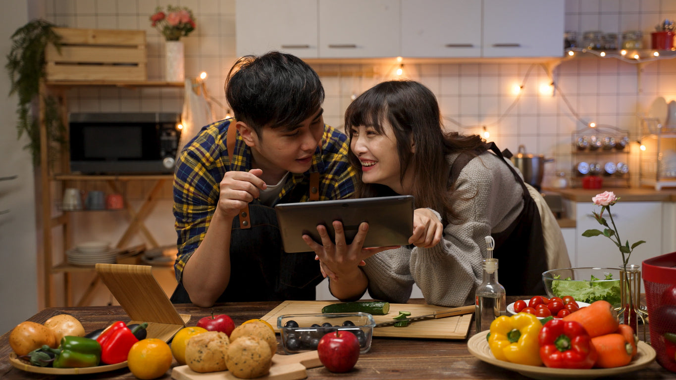A young couple preparing a low FODMAP Valentine’s Day in their kitchen. They are reading a recipe for gut friendly dinners on a tablet, talking and laughing. Their countertop is covered in fresh vegetables.