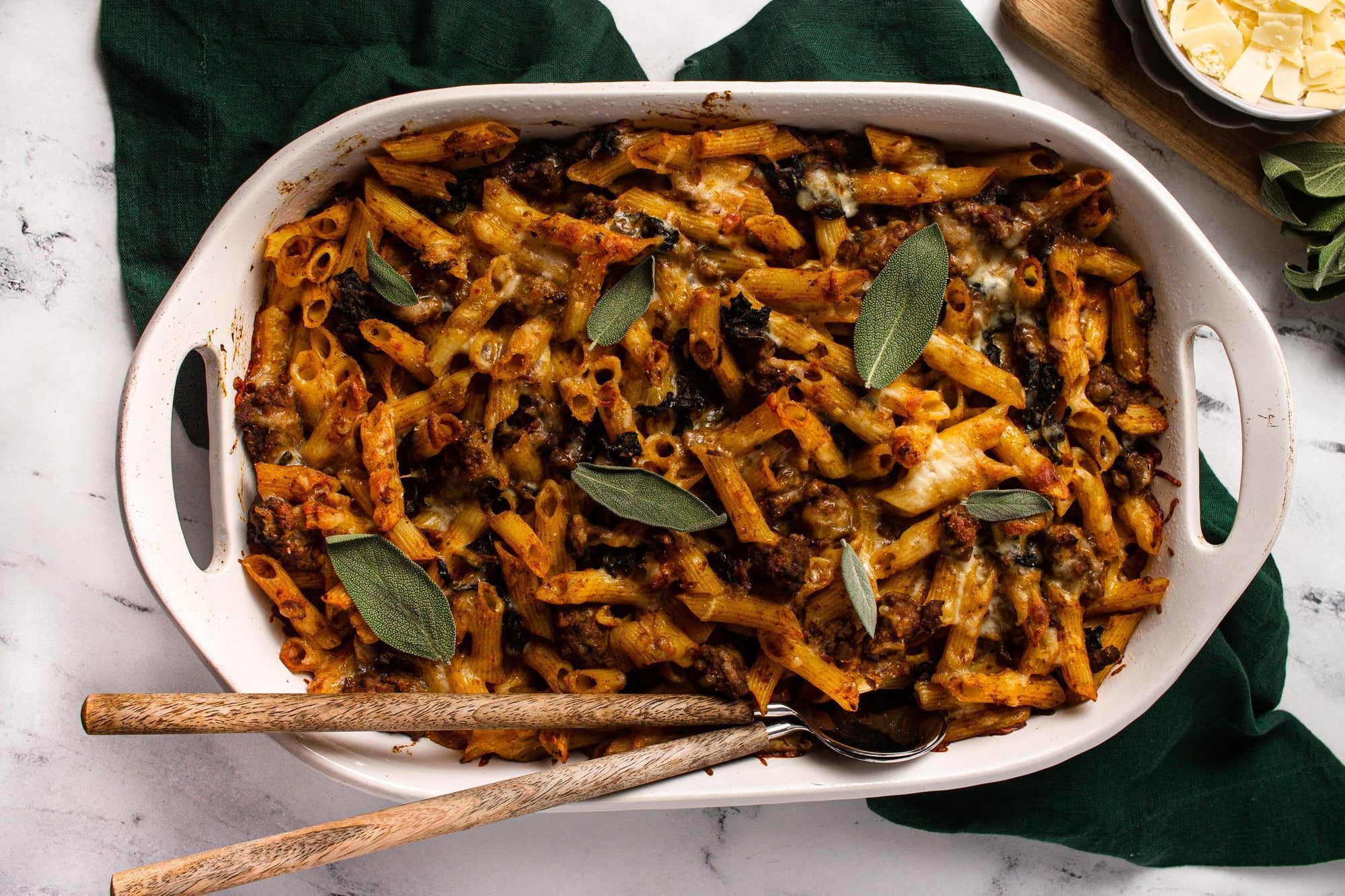 A casserole of Fody's Sausage, Kale and Pumpkin Baked Ziti. Gluten-free pasta coated with pumpkin and sausage is topped with whole sage leaves. The casserole is white, and two wooden spoons protrude from this low-fodmap pasta dish. 