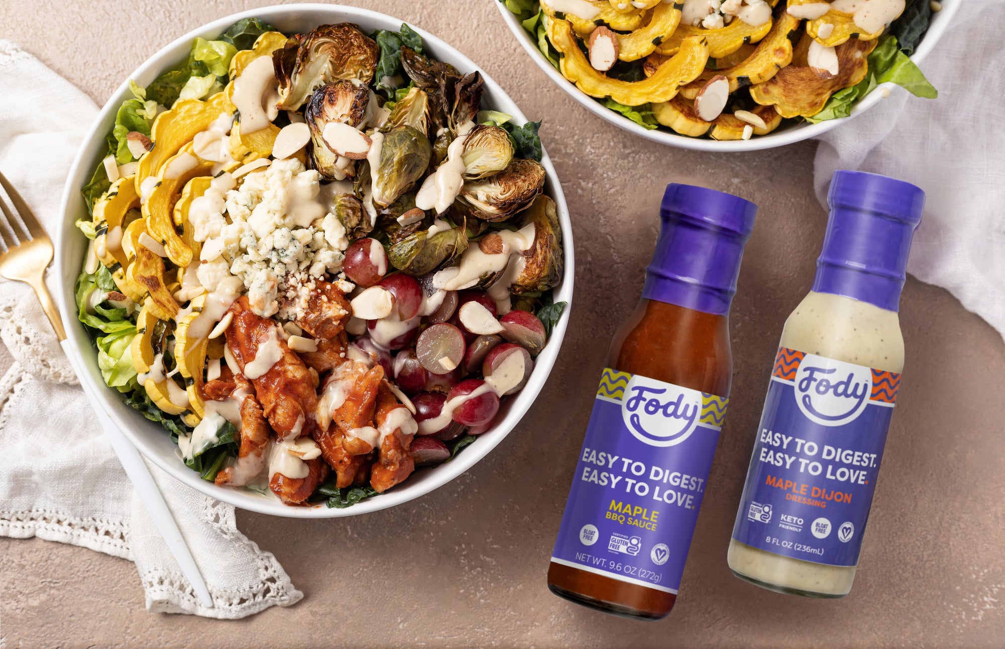 An image of Fody's Maple BBQ chicken and roasted veggie salad. Two large round bowls filled with the aforementioned ingredients sit on a wooden table beside lacy napkins and two bottles of Fody's salad dressing.