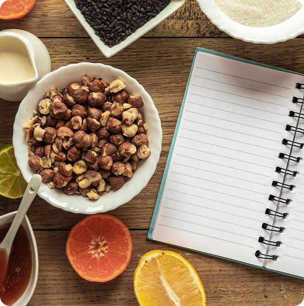 Blank notepad and pen on a wooden table beside halved oranges, berries, nuts and fresh vegetables, set up for planning midweek dinners