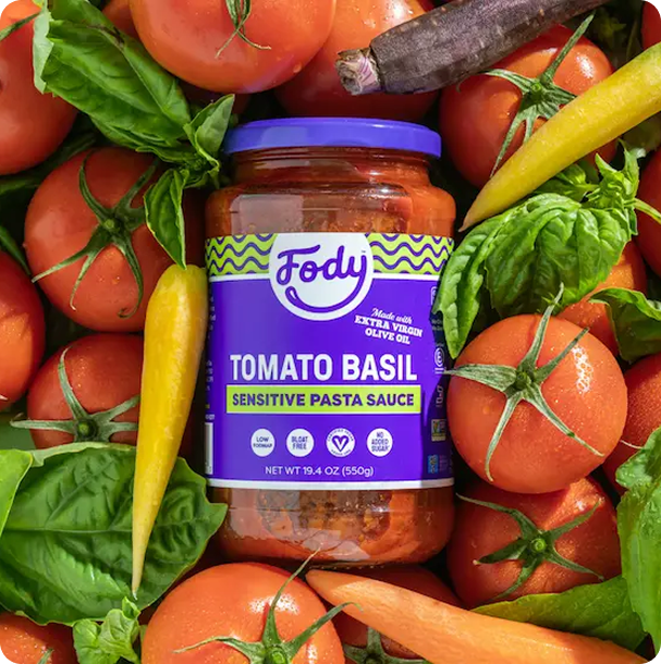 Jar of Fody Tomato Basil Pasta Sauce nestled among fresh tomatoes, rainbow carrots and greens, a simple base for a Low FODMAP meal