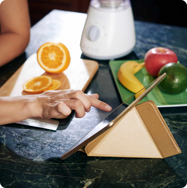 Hands writing a weekly meal plan on a notepad at a wooden kitchen table, surrounded by fresh fruit, vegetables, and a glass of water