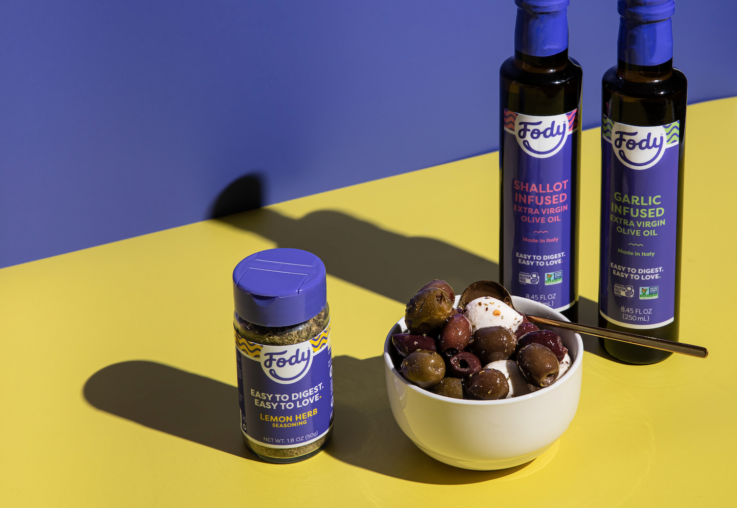 Fody Shallot Infused and Garlic Infused Olive Oils beside a Lemon Herb Seasoning bottle and a bowl of olives on a yellow and blue surface.