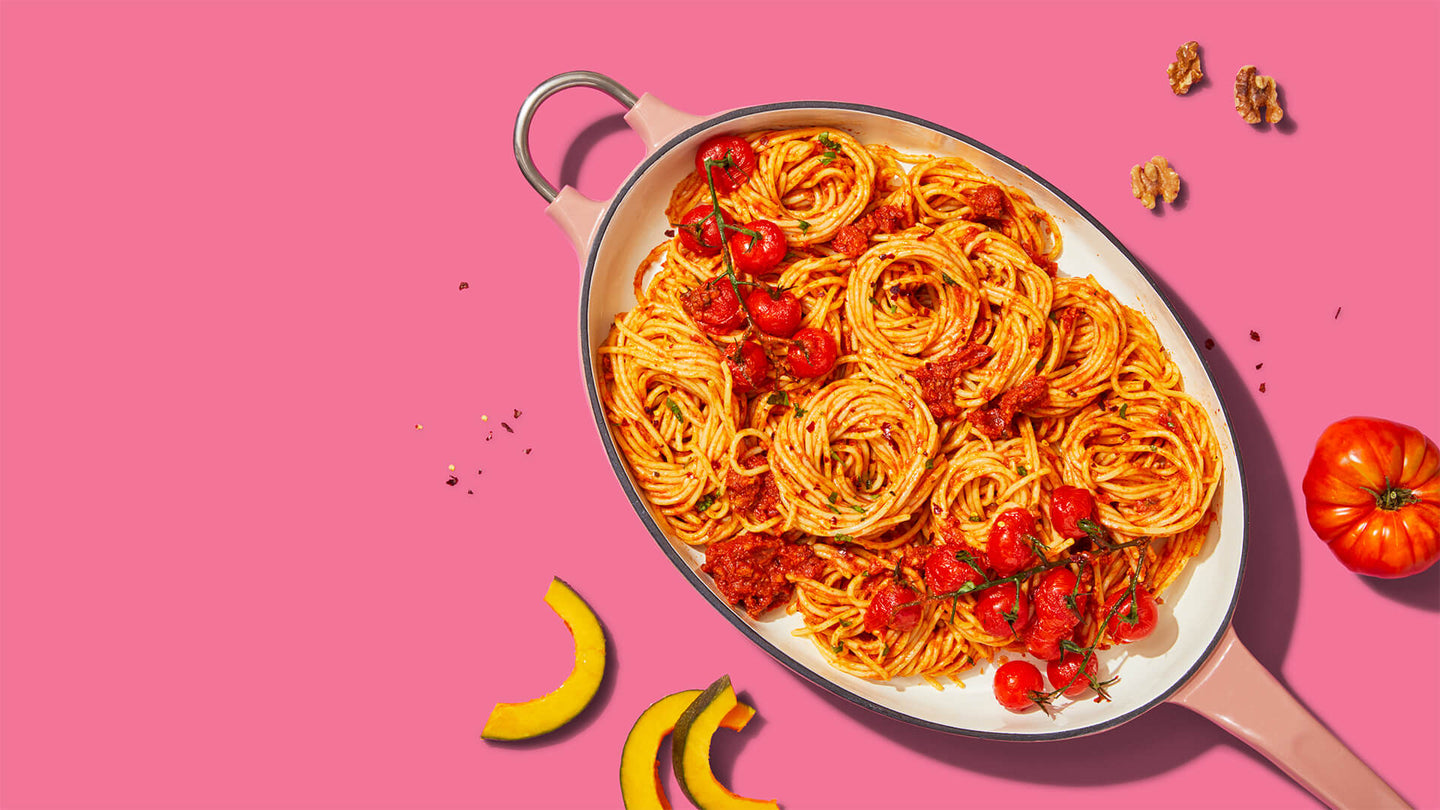 Bowl of spaghetti with tomato sauce and cherry tomatoes on a pink background, styled with yellow squash and walnuts.